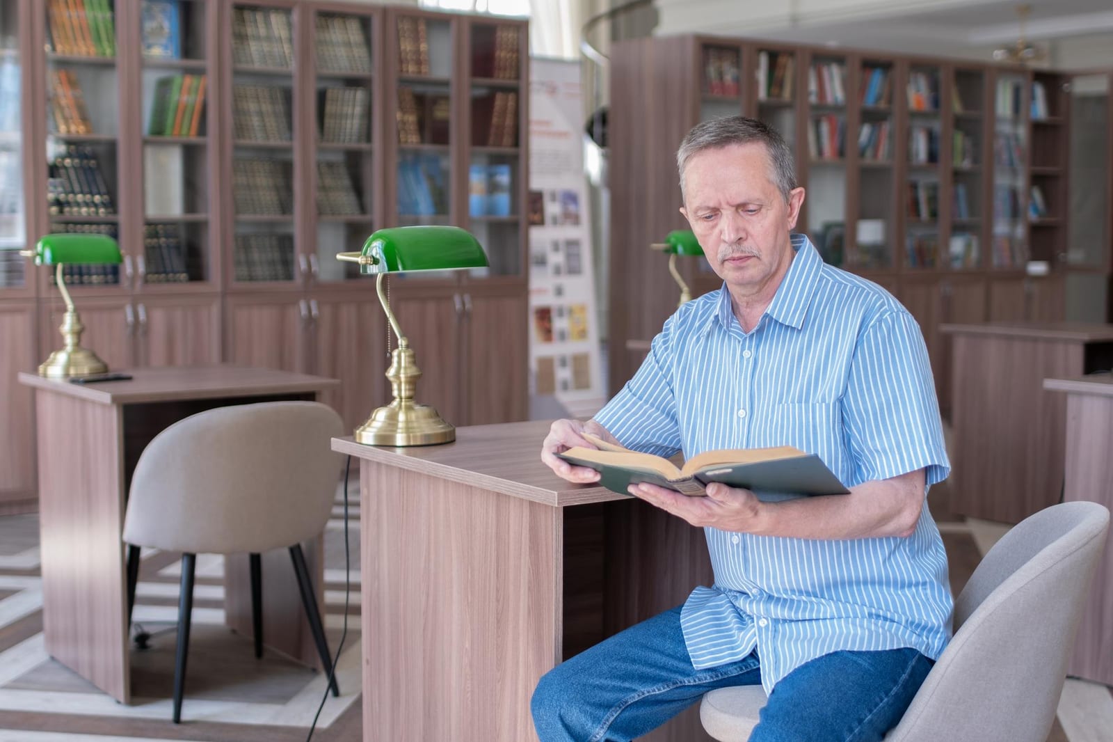 Leitor sentado a uma mesa de biblioteca, com um livro aberto nas mãos e luminárias verdes ao lado